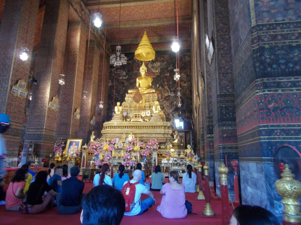 Wat Po in Bangkok. In einer thailändischen Tempelhalle sitzen Tempelbesucher und Touristen auf dem Boden, der mit Teppich ausgelegt ist, und betrachten eine vergoldete Buddhastatue, die auf einem hohen vergoldeten Altar platziert ist.