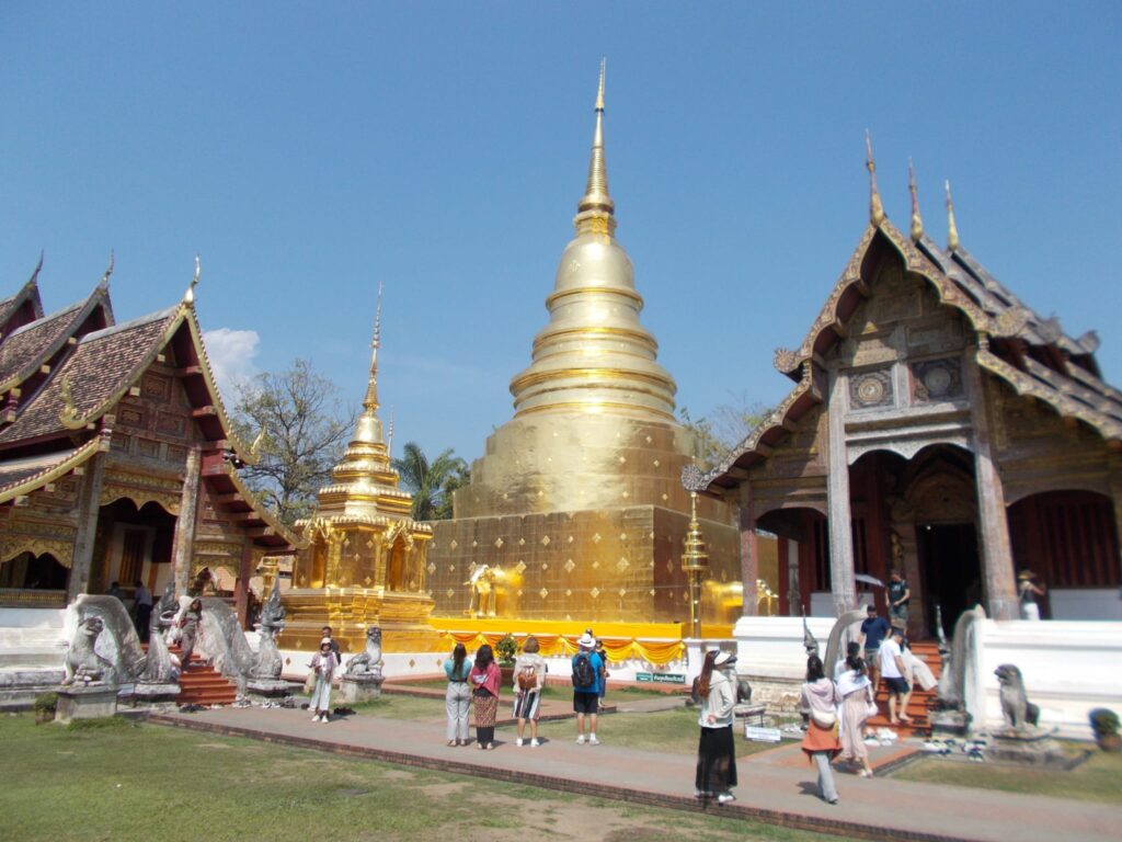 Wat Phra Singh in Chiang Mai, Thailand. Man sieht ein Tempelgelände mit zwei kleinen Hallen, in der Mitte eine vergoldete Stupa, davor Touristen, die sie betrachten. 
